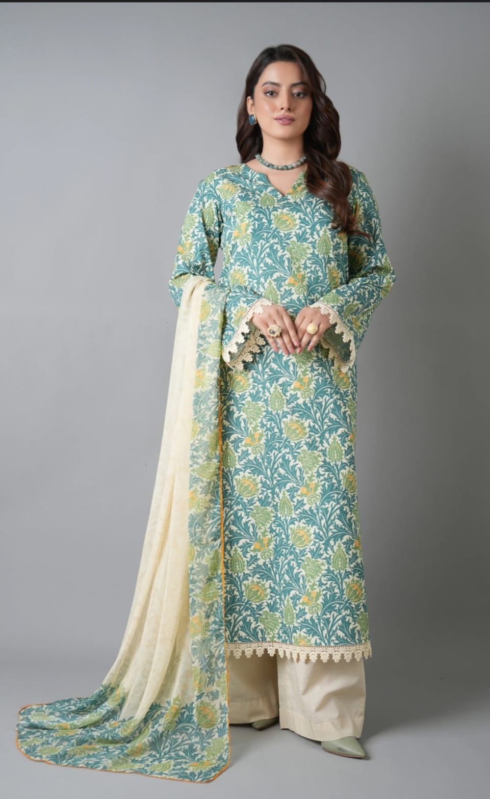 Woman wearing a teal floral traditional outfit with a beige dupatta on a gray background