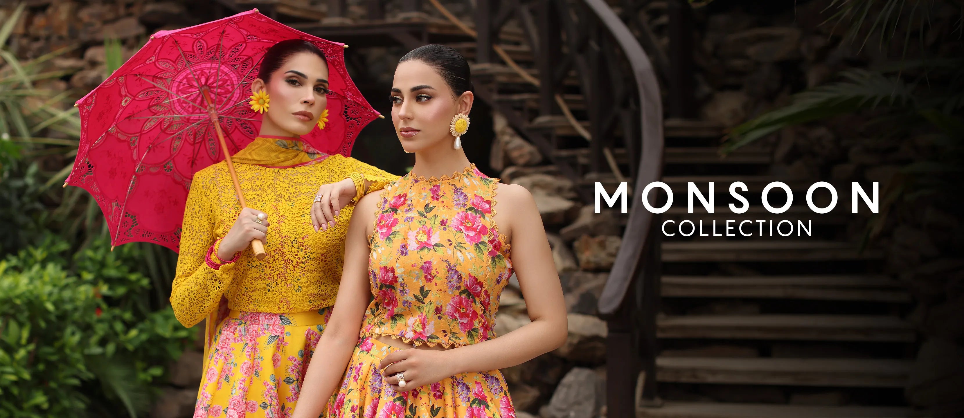 Two women in colorful monsoon outfits with a pink umbrella, standing in front of a rustic background with the 'Monsoon Collection' text.