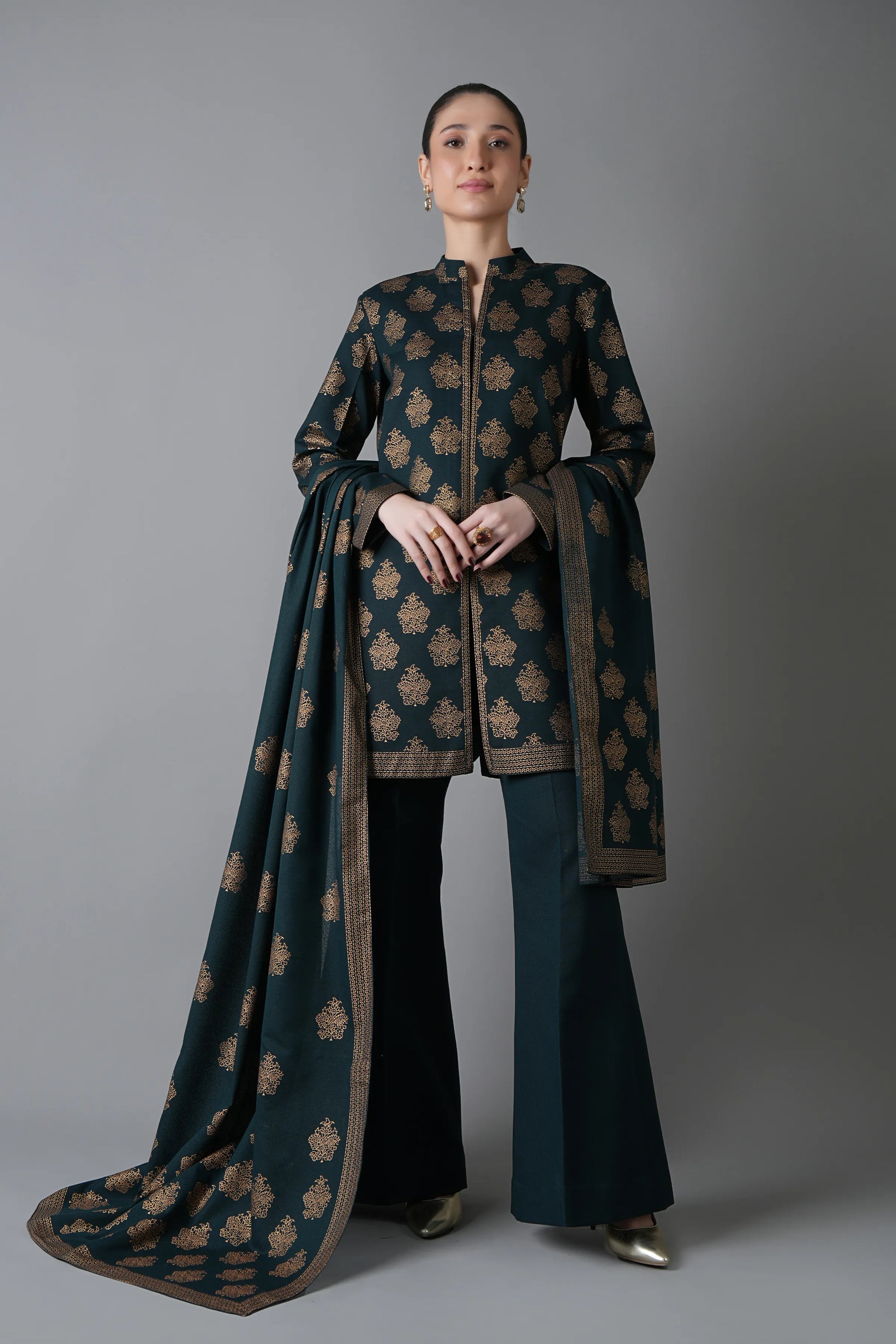 Woman wearing a traditional outfit with a long dark green and brown patterned scarf against a gray background