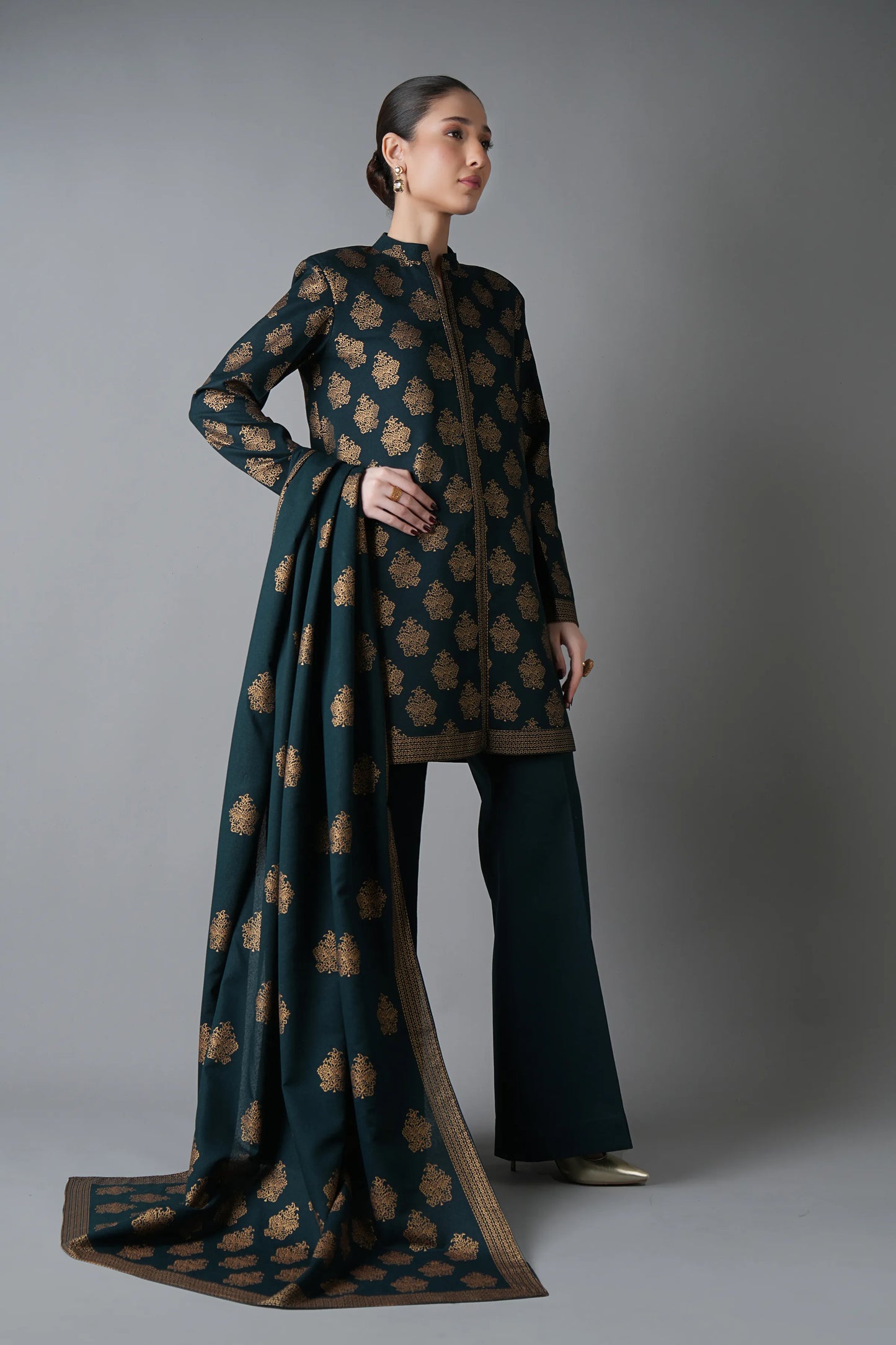 Woman wearing a dark green traditional outfit with a patterned dupatta on a gray background