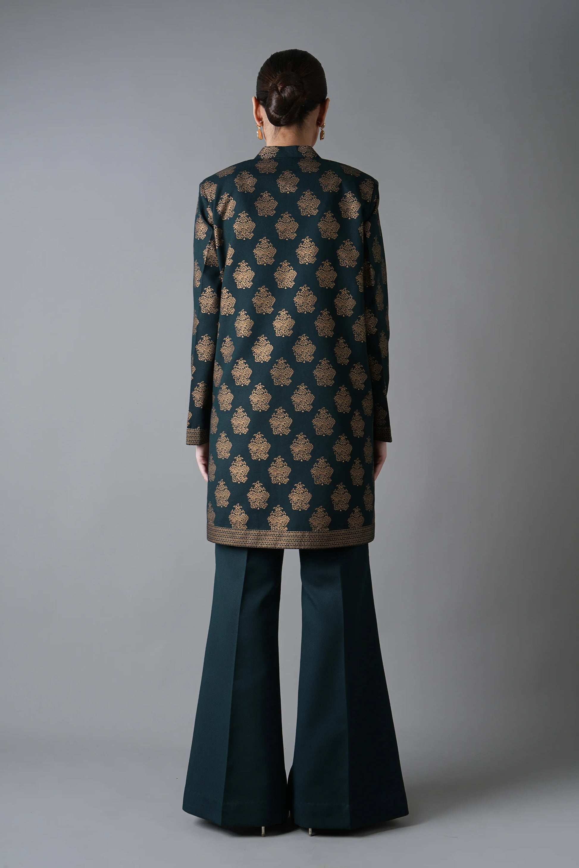 Person wearing a patterned coat and dark green flared pants against a gray background