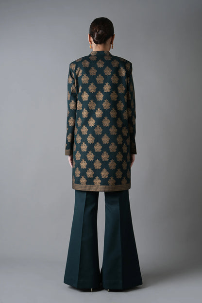 Person wearing a patterned coat and dark green flared pants against a gray background